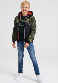 Vaatteet, jossa on pehmustettu takki, jossa on camo-vihreä ja musta muotoilu, oranssi vuori, huppu ja valkoiset yksityiskohdat; yhdistetty sinisiin farkkuihin ja valkoisiin tennareihin.