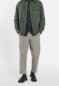 Groen overhemd met knopen en twee borstzakken, marineblauw shirt eronder, beige broek en zwarte instappers. Casual fit met opgerolde mouwen.
