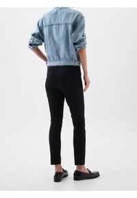 Personne debout, portant une veste en jean bleu clair avec les manches retroussées, un pantalon slim noir et des mocassins noirs sur fond blanc.