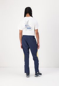 Marine joggingbroek met gele zijpanelen, elastische tailleband; wit T-shirt met een blauwe bootprint op de achterkant. Draagt bij zwarte sneakers.