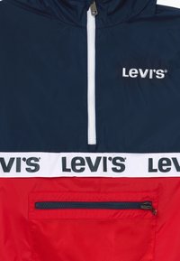 Röd och marinblå vindjacka med en ficka med dragkedja, vit dragkedja och randig midja med "Levi's"-logotypen i svart. Smidigt, lätt material.