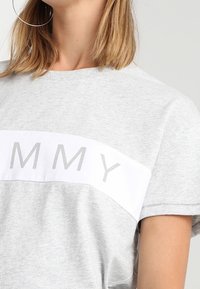 Grå t-shirt i bomull med ett vitt band som har texten "TOMMY" i grå bokstäver. Kort ärm och en avslappnad passform.