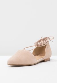 Ballerine nude in suede, con punta affusolata, lacci alla caviglia e tacco basso. Il design presenta delle aperture sui lati e un piccolo dettaglio decorativo.