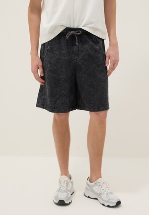 Hombre vistiendo pantalones cortos casuales negros con cordón, zapatillas blancas y una camiseta blanca frente a un fondo claro y liso.