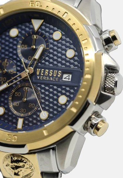 Montre avec un cadran texturé bleu et des accents dorés, boîtier en acier inoxydable argenté, trois sous-cadrans et emblème de lion sur le bracelet. Comprend une fenêtre de date.