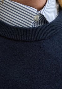 Suéter de color navy con cuello acanalado, superpuesto a una camisa de cuello con rayas azul claro y blanca, que presenta cierre de botones y una textura de tela suave.