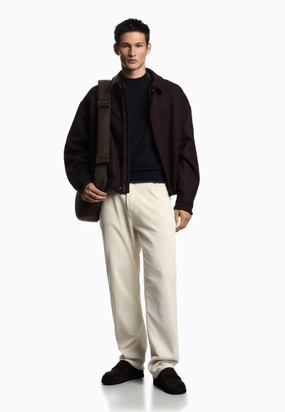 Una modella indossa una giacca zip di colore marrone scuro sopra un maglione nero, pantaloni di velluto a coste color crema e pantofole marroni, mentre porta una borsa di colore verde scuro.