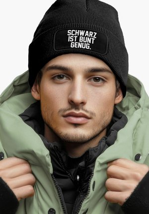 Black ribbed beanie with white text "SCHWARZ IST BUNT GENUG" worn with a green puffy jacket featuring a zip closure and black inner lining.