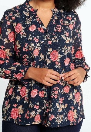 Nainen napittaa laivastonsinisen puseron, jossa on vaaleanpunainen ja beige kukkakuosi sekä pienet valkoiset pilkut, pitkät hihat ja röyhelöidyt kalvosimet.