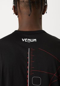 Černé tričko s bílým textem "VENUM" na zadní straně, s grafickými prvky v červené a bílé barvě a rovně střiženým lemem.