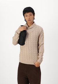 Un pull en maille beige avec un col rabattu, associé à un pantalon en velours côtelé marron foncé. Le modèle tient un sac à bandoulière noir.