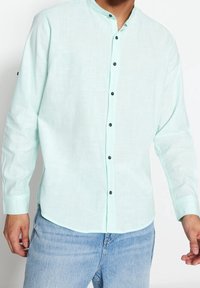 Camicia a maniche lunghe verde menta chiaro con colletto rotondo, bottoni neri e un tessuto strutturato. Finitura liscia e orlo curvato.