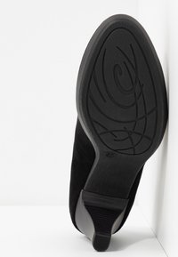 Chaussure à talons en daim noir pour femmes, positionnée semelle vers le haut contre un coin blanc, montrant la semelle en caoutchouc texturée en détail et le talon carré.