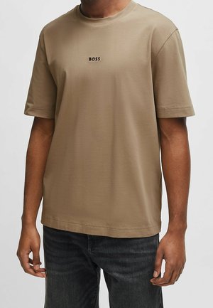 Homme portant un t-shirt marron clair uni à manches courtes avec le mot "BOSS" imprimé en petites lettres noires sur la poitrine et un jean foncé.