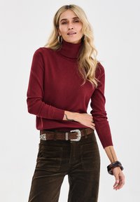 Frau mit langen blonden Haaren, die einen roten Rollkragenpullover, braune Cordhosen, einen Gürtel mit silberner Schnalle, Creolen und Armbänder trägt.