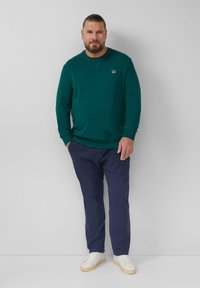 Ciemnozielony sweter z okrągłym dekoltem, z małym logo. W zestawie z granatowymi spodniami i białymi trampkami. Gładka tekstura.