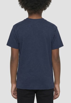 Niño con cabello corto y rizado, vistiendo una camiseta azul marino lisa de manga corta y pantalones negros, de pie de espaldas sobre un fondo claro.