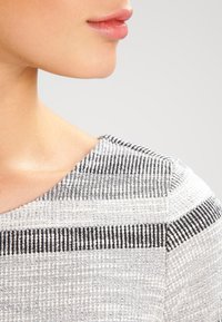 Detailní záběr na texturovanou látku s horizontálními pruhy v bílé, černé a šedé, s hladkým výstřihem a přirozeným tónem pleti v pozadí.