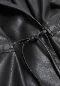 Gros plan sur un vêtement en cuir noir avec une lanière en cuir nouée et des coutures texturées visibles sur un matériau lisse.