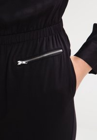 Combinaison noire avec une taille élastique, poche latérale avec une fermeture éclair argentée, et manches longues au toucher lisse.