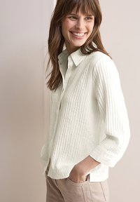 Camicia bianca a bottoni, con un collo classico, vestibilità leggermente ampia e maniche a tre quarti. Indossata con pantaloni chiari.