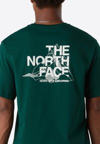 Person iført en mørkegrøn t-shirt med en hvid bjerggrafik og teksten "THE NORTH FACE NEVER STOP EXPLORING" på ryggen.