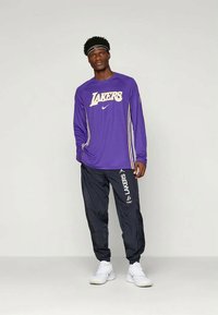 Lila hosszú ujjú póló, rajta "LAKERS" felirattal és Nike logóval, fekete nadrággal, amelyen a "LAKERS" logó található, fehér tornacipővel viselve.