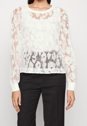 Kvinde iført hvid gennemsigtig bluse med blomsterprint over hvid top, kombineret med sorte bukser med højtaljet pasform, stående foran en ensfarvet baggrund.
