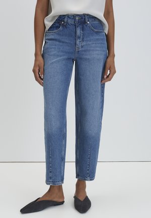 Persoon die mid-rise rechte blauwe spijkerbroek, wit topje en zwarte slip-on puntige platte schoenen draagt, staand tegen een effen achtergrond.