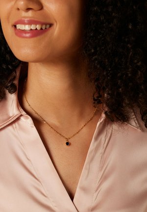 Goldene Kettenhalskette mit einem kleinen, runden, dunkelblauen Anhänger, getragen über einer hellpinkfarbenen Satinbluse. Hat ein schlichtes, elegantes Design.