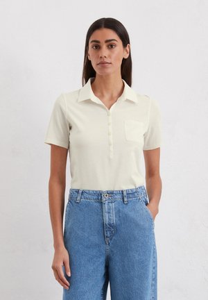 Kurzärmliges, kragenloses Poloshirt in hellbeige mit Knopfleiste und Brusttasche, kombiniert mit hoch taillierten blauen Jeans.
