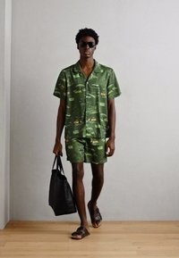 Grønt sæt med kortærmet skjorte og shorts med grafisk print, parret med sorte sandaler og en tote bag, mod en neutral baggrund.