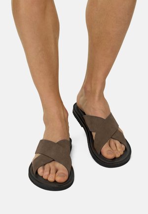 Piernas masculinas con sandalias de ante marrón con tiras anchas cruzadas y suelas negras, sobre un fondo blanco.