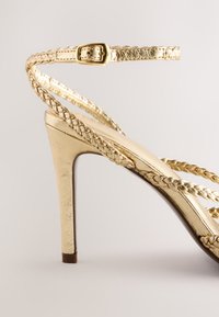 Sandale à talons hauts dorée avec des brides tressées et un dos fermé. Présente un design élégant et une finition métallique brillante.