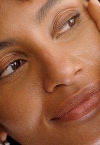Gros plan sur un visage avec une peau lisse et aux tons chauds, des lèvres pulpeuses et des yeux brun foncé. Les caractéristiques remarquables incluent des pommettes bien définies et un maquillage naturel.