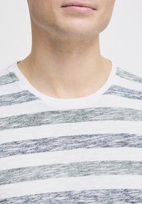 T-shirt rayé en blanc, présentant des rayures horizontales inégales dans des tons de vert et de bleu, avec un col rond côtelé et une texture douce.