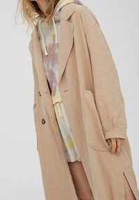 Abrigo oversized de lino beige con grandes bolsillos, cierre de un solo botón y corte relajado. Lleva puesto sobre una sudadera con capucha de tie-dye en tonos pastel.