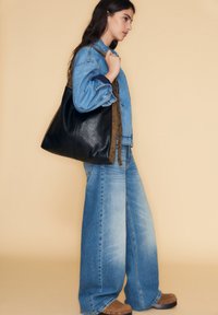 Svart läder tote-väska med en mörkbrun mockaband. Modellen bär en blå denimjacka och vida jeans, med bruna träskor. Neutral bakgrund.