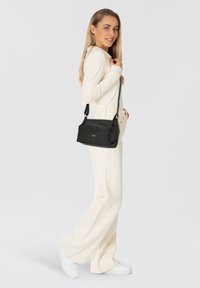 Borsa a tracolla in pelle nera con forma strutturata, spallaccio regolabile e discreto logo. Modella che indossa un top crema e pantaloni a gamba larga.