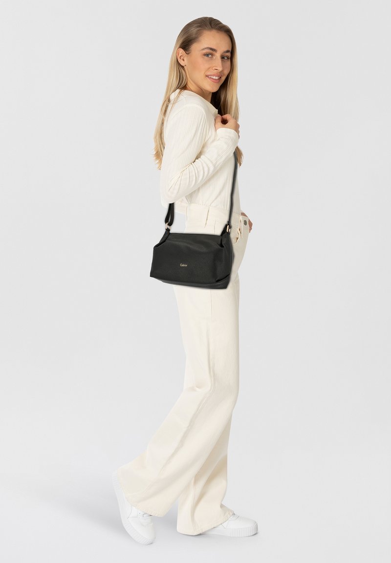 Borsa a tracolla in pelle nera con forma strutturata, spallaccio regolabile e discreto logo. Modella che indossa un top crema e pantaloni a gamba larga.