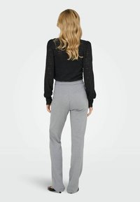 Femme aux longs cheveux blonds portant un blouse noire à manches longues et un pantalon gris taille haute, se tenant de dos contre un fond uni.