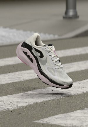 Λευκό και μαύρο αθλητικό παπούτσι Nike με ροζ σόλα στον αέρα πάνω από διάβαση πεζών σε δρόμο, με χαλίκι και μεταλλικό πόλο στο φόντο.