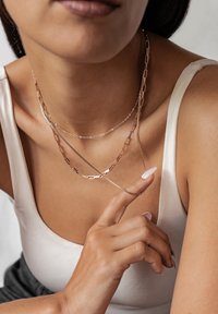 Trzy warstwowe naszyjniki z srebrnymi i różowo-złotymi łańcuszkami z prostokątnymi ogniwami, noszone na szyi modelki. Widoczna gładka skóra i neutralna bluzka.
