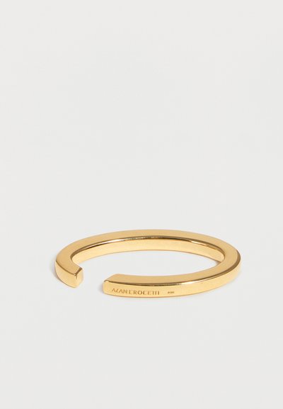 Χρυσό ανοιχτό βραχιόλι με λεία επιφάνεια και χαραγμένη τη λέξη «ALAN EROCETTI» στη μία πλευρά.