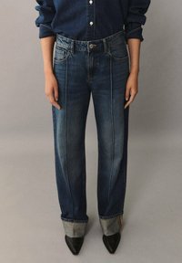 Vaqueros de mezclilla de corte holgado, color azul, con costuras visibles y puños enrollados, combinados con zapatos negros de punta.