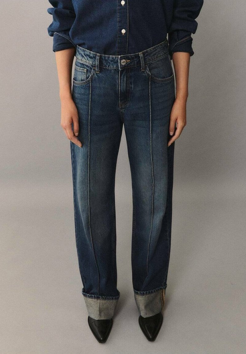 Vaqueros de mezclilla de corte holgado, color azul, con costuras visibles y puños enrollados, combinados con zapatos negros de punta.