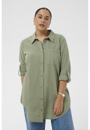 Kobieta z zaczesanymi do tyłu włosami, ubrana w zieloną koszulę na guziki z podwiniętymi rękawami oraz niebieskie jeansy, stojąca na jednolitym białym tle.