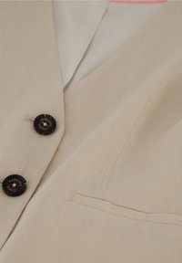 Beige Blazer aus strukturiertem Stoff, zwei schwarze Knöpfe mit Logodetail und einer Brusttasche. Innen mit einem weißen Stoff gefüttert.