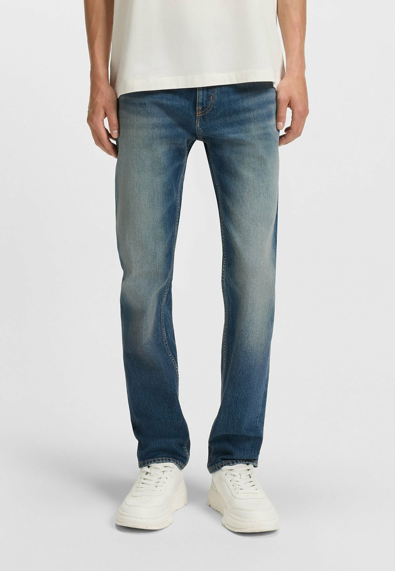 Blue denim jeans featuring a straight-leg cut, fading details, and an orange stitching accent, paired with white sneakers.