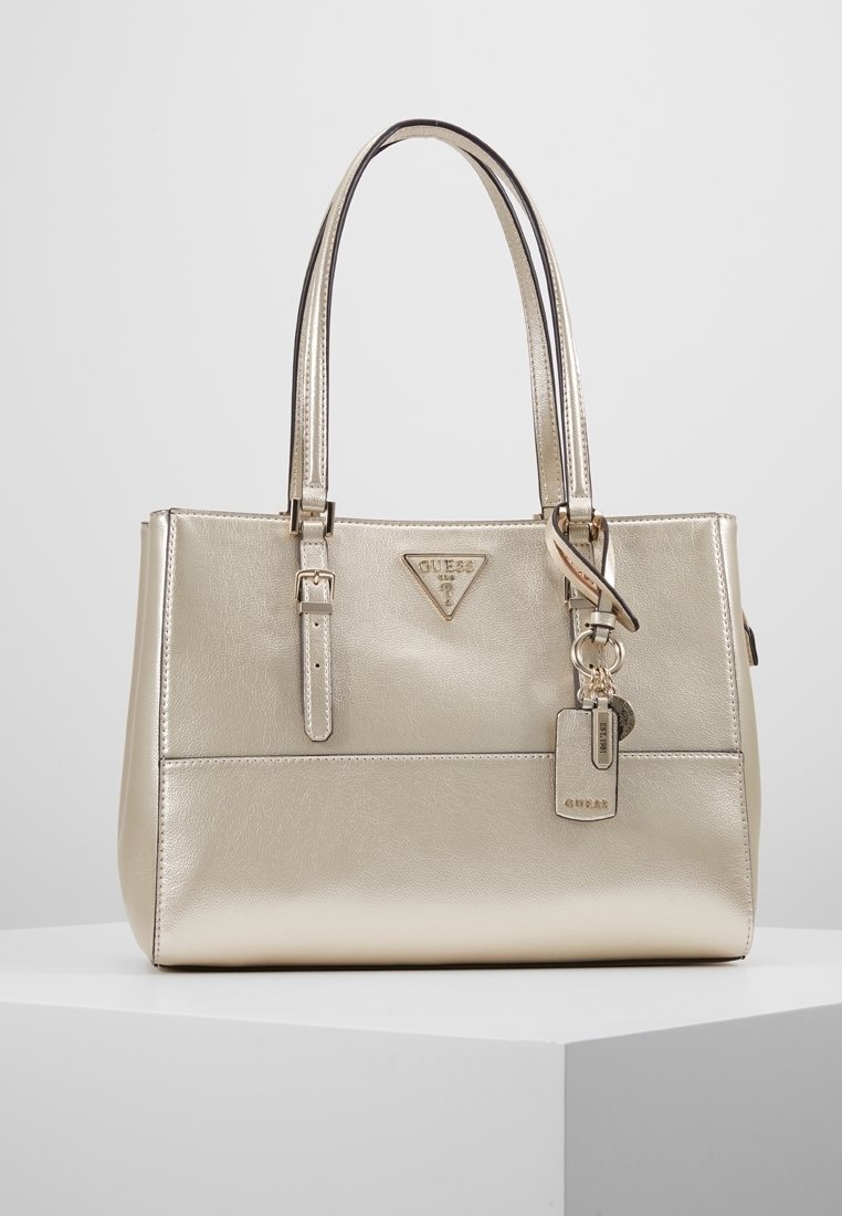 Kultainen keinonahkainen olkalaukku, jossa on jämäkkä muoto, kaksinkertaiset kahvat, metalliosat ja nimilappu. Laukussa on edessä logoyksityiskohta.